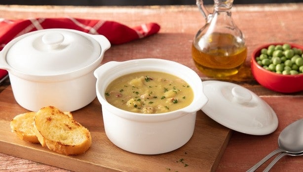 Tigela de sopa de ervilha com pedaços de bacon e cheiro-verde, acompanhada de torradas, azeite e ervilhas frescas ao fundo.