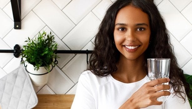 Mulher negra de cabelos longos ondulados está segurando um copo de água para responder a pergunta se suco substitui água.
