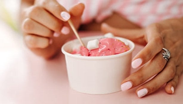 Mãos de uma mulher segurando um pote branco de sorvete com a cor rosa. A imagem representa a dúvida se tomar sorvete gripado 