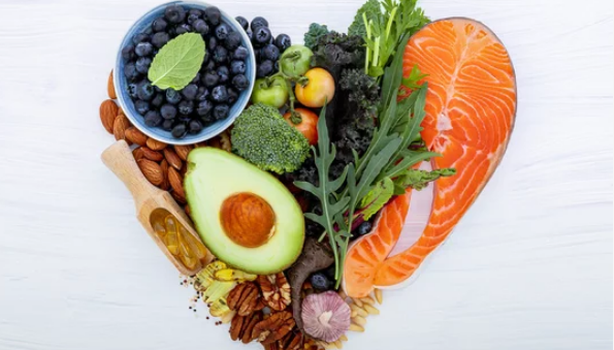 Imagem de alimentos saudáveis dispostos em forma de coração, incluindo salmão, abacate, mirtilos e nozes sobre fundo branco.