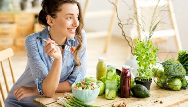 Mulher sorrindo à mesa com salada e sucos naturais, simbolizando escolhas saudáveis e consciência alimentar.