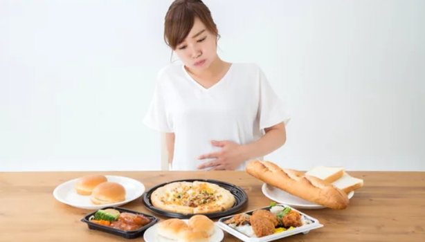 Mulher sentada à mesa com vários pratos de comida, segurando a barriga, representando saciedade.