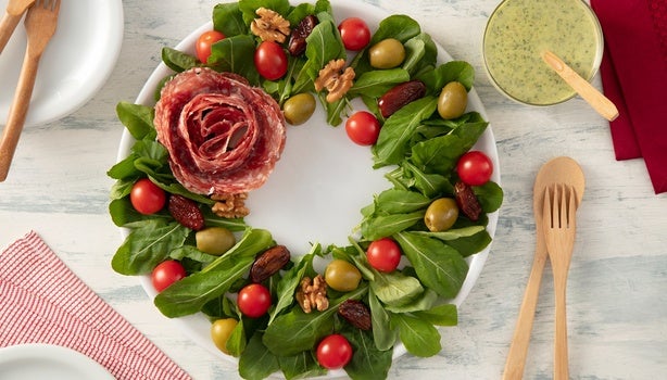 Guirlanda de salada com folhas verdes, tomatinhos, azeitonas, nozes, tâmaras e flor de salame ao centro.