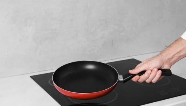 Mão segurando frigideira antiaderente sobre cooktop de indução, pronta para fazer a cura da panela.