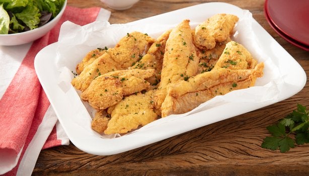 Tiras de frango empanado com ervas, servidas em travessa branca sobre mesa de madeira.