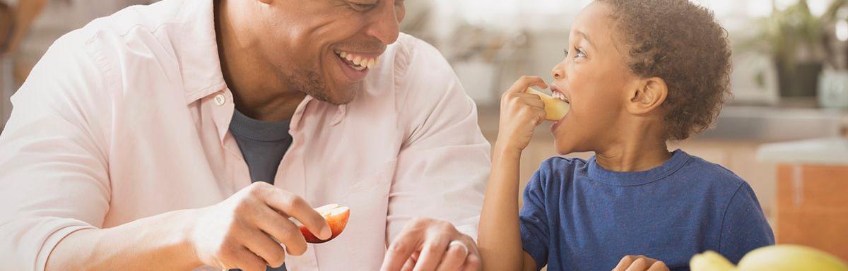 Lanche da Tarde: Um homem negro com uma maçã na mão e uma criança negra comendo uma maçã 