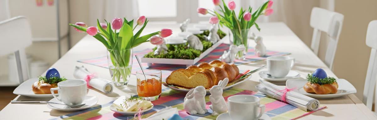Decoração de Páscoa: Mesa decorada com pratos, talheres, pães, vaso com flores e xícaras