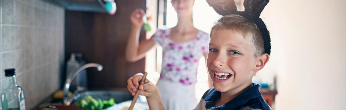 Sexta-Feira Santa: Menino com tiara de coelho sorrindo enquanto segura uma colher ao lado da mãe na cozinha