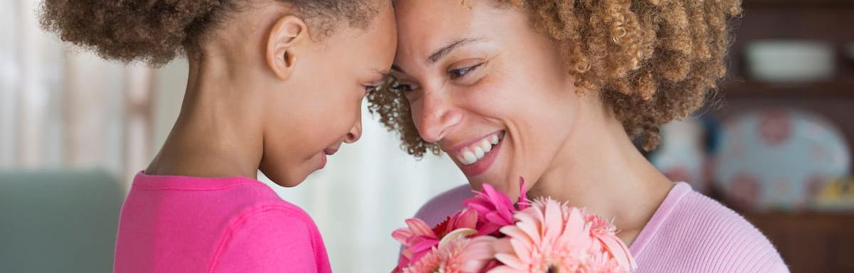 Dia das Mães: Mãe e filha bem próximas encostando a testa uma na outra, sorrindo com uma flor no meio