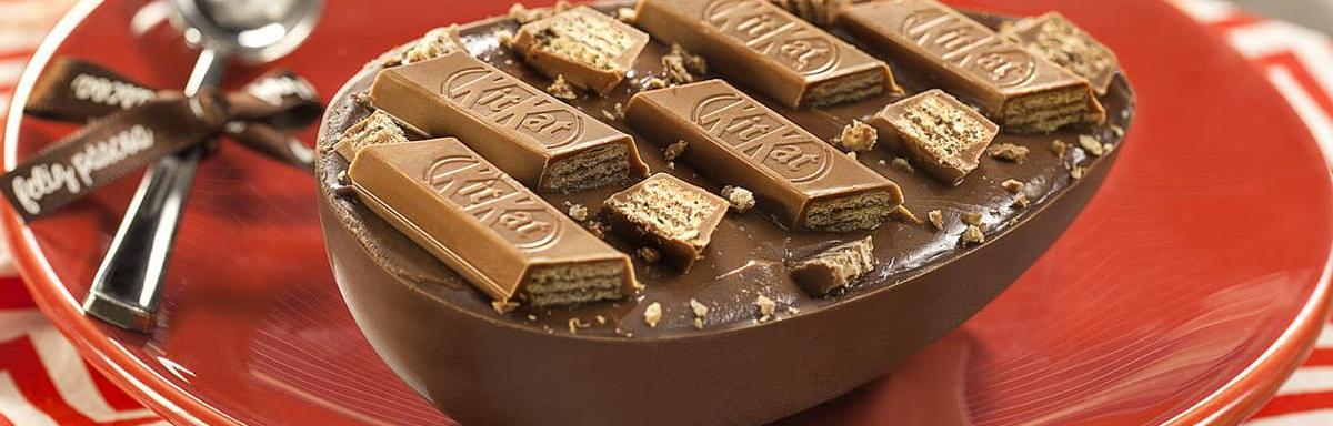 Ovo de colher: Ovo de páscoa com pedaços de chocolate KitKat em cima, em um prato ao lado de uma colher