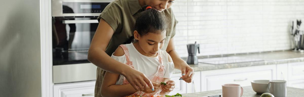 Almoço de Dia das Mães: Mulher negra adulta na cozinha ajudando criança negra a cozinhar
