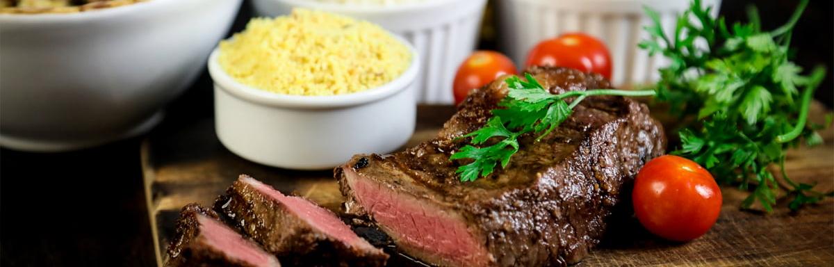 Carne cortada em três pedaços com tomates em volta, algumas tigelas, em cima de uma mesa de madeira