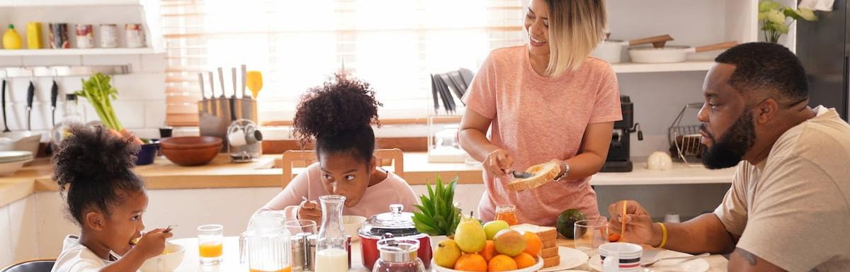 Dicas de café da manhã:Duas crianças negras à esquerda, dois adultos negros à direita, com frutas, leite, pão e café na mesa