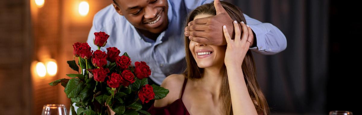 Jantar para o Dia dos Namorados: Homem negro segurando um buquê de flores e com a mão nos olhos de uma mulher branca 