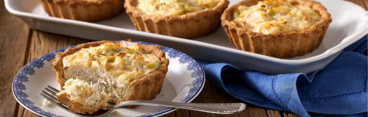 Quiche de alho-poró sendo cortado sobre um prato com um garfo ao lado. Ao fundo, mais 3 quiches inteiros