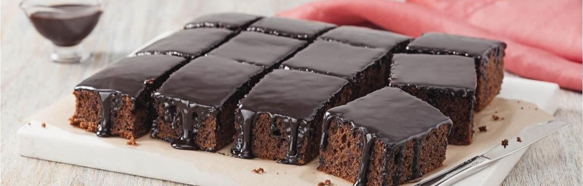 Prato com um bolo de chocolate cortado em pedaços quadrados e uma faca, ao fundo um pano vermelho e um potinho com chocolate 