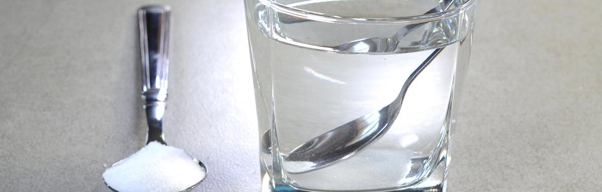 Copo com água e colher dentro, ao lado de colher com sal sobre uma mesa clara, representando soro caseiro.