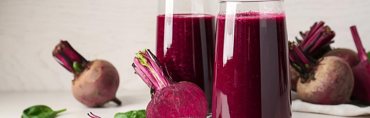 Copos com suco de beterraba, rodeados por beterrabas frescas cortadas e folhas verdes sobre superfície clara.