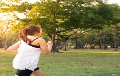 Imagem de uma mulher correndo em um parque arborizado no final da tarde.