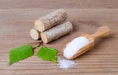Em uma mesa, há três madeiras empilhadas com duas folhas verdes à frente. Ao lado, temos o xilitol, substituto do açúcar. 