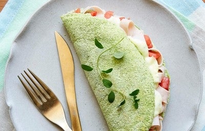Imagem de uma tapioca verde de espinafre recheada com queijo e tomate sobre um prato cinza.