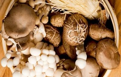 Em uma mesa de madeira, há uma cesta de palha com diversos tipos de cogumelos comestíveis.