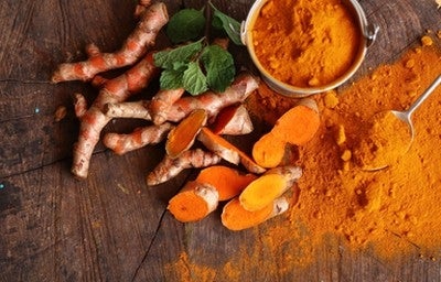 Raízes de cúrcuma frescas cortadas e em pó, dispostas sobre mesa de madeira rústica com folhas verdes decorativas.