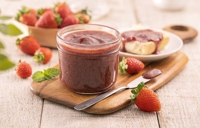 Pote de geleia de morango sobre tábua de madeira, com morangos frescos e pão com geleia ao fundo.