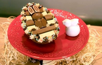 Ovo de Páscoa recheado e decorado com chocolates KitKat e confeitos coloridos, servido em prato vermelho com coelho branco.