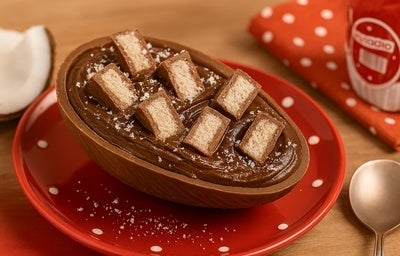 Ovo de chocolate recheado com creme e decorado com pedaços de prestígio, servido em prato vermelho com poás brancos.