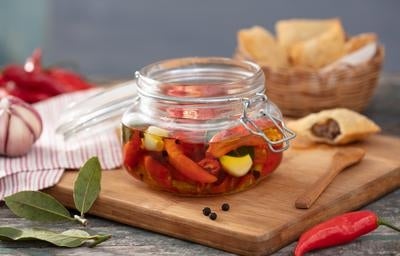 Pimenta em conserva caseira com alho e azeite em pote de vidro sobre tábua de madeira.