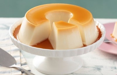 Pudim de leite condensado em forma redonda com calda de caramelo, servido em prato branco sobre mesa clara. 