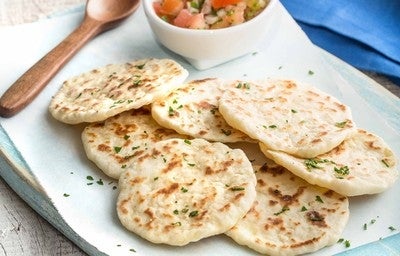 Imagem de pães achatados dourados servidos em prato branco, decorados com salsa e acompanhados de molho em tigela.