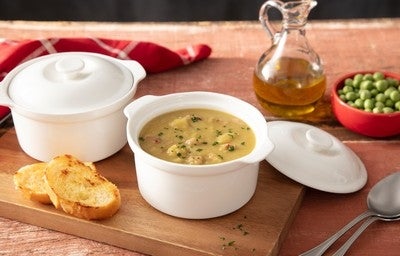 Tigela de sopa de ervilha com pedaços de bacon e cheiro-verde, acompanhada de torradas, azeite e ervilhas frescas ao fundo.