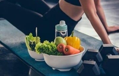 Mulher com roupas de ginástica sentada em um tapete no chão da academia. Ao lado, uma tigela com legumes e Halteres.