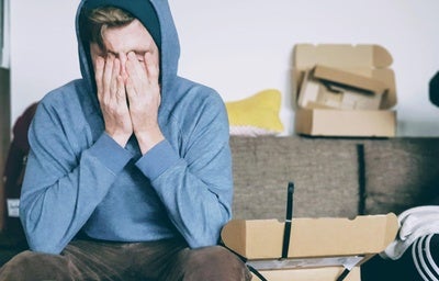 Cortisol: o que é e como funciona o hormônio do estresse! Homem sentado em seu sofá, rodeado de caixas abertas. Ele usa um casaco azul de touca e coloca as mãos no rosto, preocupado.