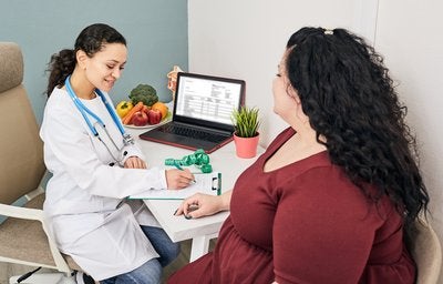 7 mitos e verdades sobre a obesidade Em um consultório, uma médica está atendendo uma mulher obesa. Ambas estão sentadas em volta de uma mesa, conversando.
