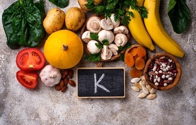 Alguns alimentos ricos em potássio, junto a um quadro negro com a letra K desenhada, representando potássio para que serve.