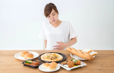 Mulher sentada à mesa com vários pratos de comida, segurando a barriga, representando saciedade.