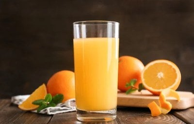Copo de suco de laranja natural sobre mesa rústica, com laranjas inteiras e cortadas ao fundo.