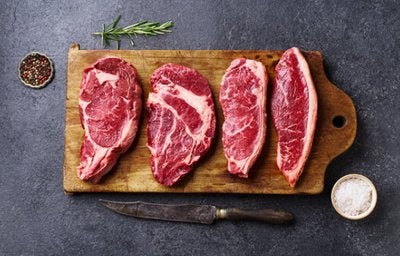 Cortes de carne bovina crua dispostos sobre tábua de madeira, com faca, sal grosso e alecrim, representando carnes para bife.