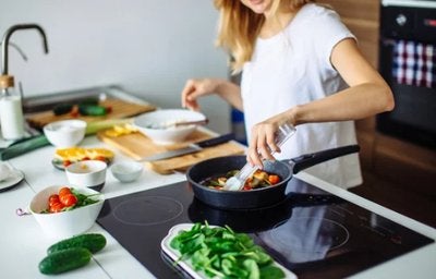 Mulher cozinhando em frigideira sobre cooktop, com ingredientes frescos organizados na bancada da cozinha.