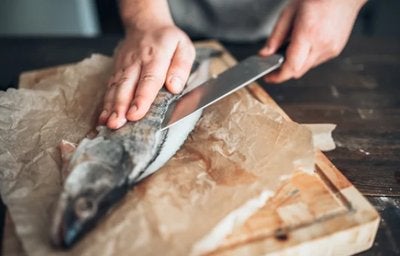 Mãos cortando peixe fresco com faca sobre tábua de madeira, preparando o alimento para o cozimento.