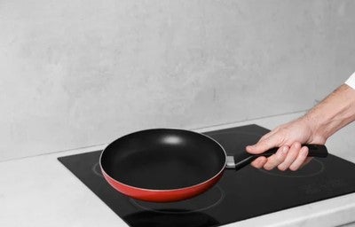 Mão segurando frigideira antiaderente sobre cooktop de indução, pronta para fazer a cura da panela.