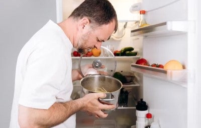Homem observa panela com comida da geladeira aberta, observando ingredientes e recipientes nas prateleiras internas.