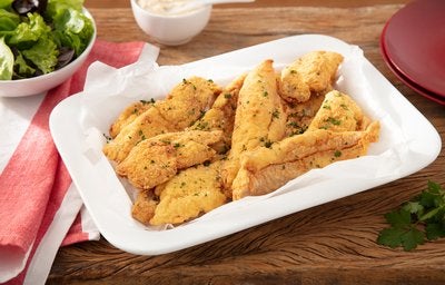 Tiras de frango empanado com ervas, servidas em travessa branca sobre mesa de madeira.