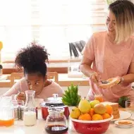Dicas de café da manhã: Duas crianças negras à esquerda, dois adultos negros à direita, com frutas, leite, pão e café na mesa