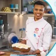 Imagem de um rapaz com uniforme branco com um bolo retangular coberto com um creme os dizeres com o nome do curso e os logos