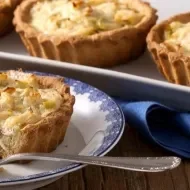 Quiche de alho-poró sendo cortado sobre um prato com um garfo ao lado. Ao fundo, mais 3 quiches inteiros