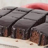 Prato com um bolo de chocolate cortado em pedaços quadrados e uma faca, ao fundo um pano vermelho e um potinho com chocolate 